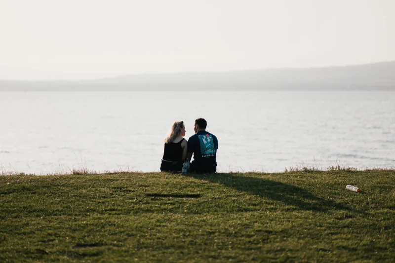 a couple sits on a grassy hill overlooking a body of water