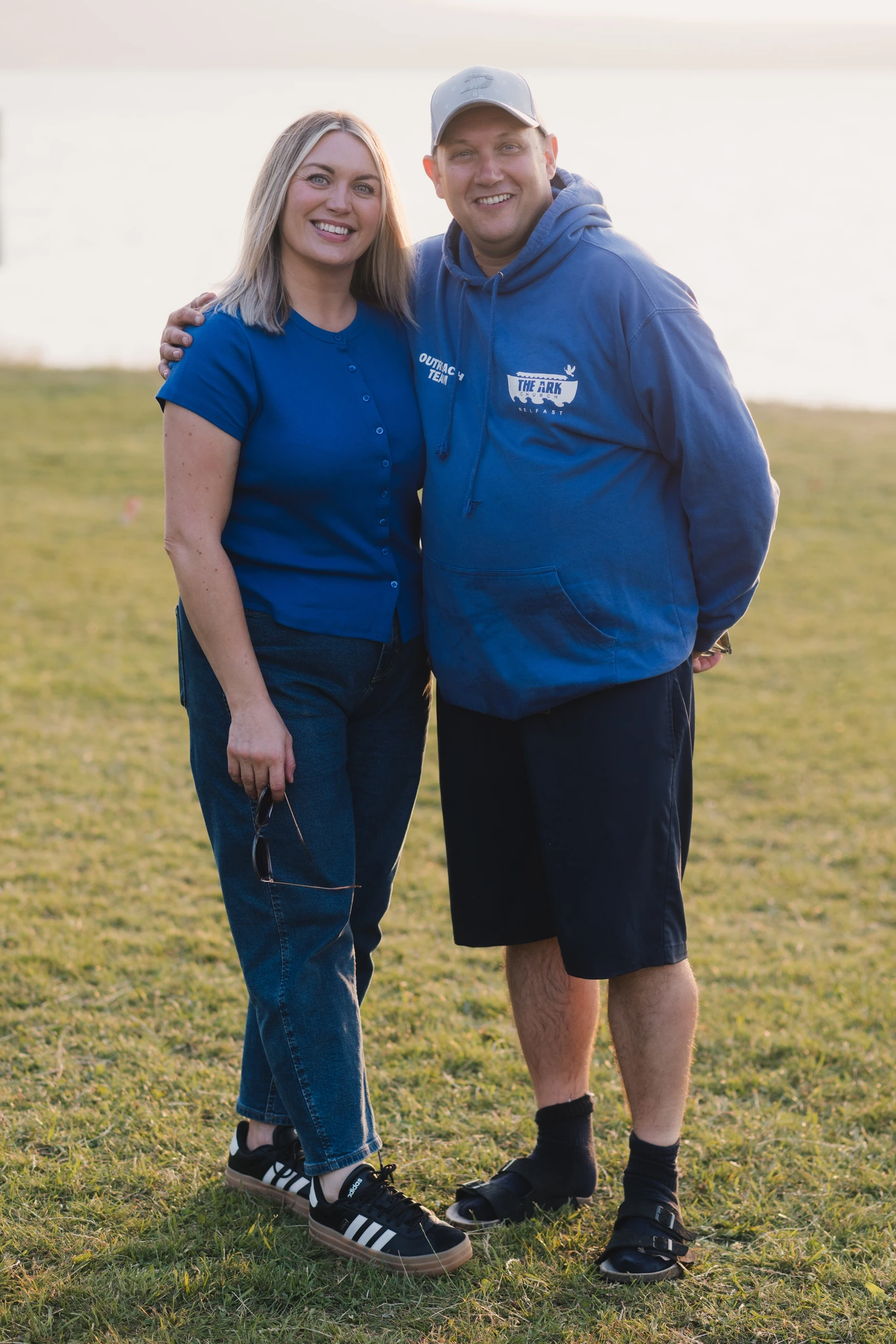 a man and woman standing on a grassy field with a lake in the background