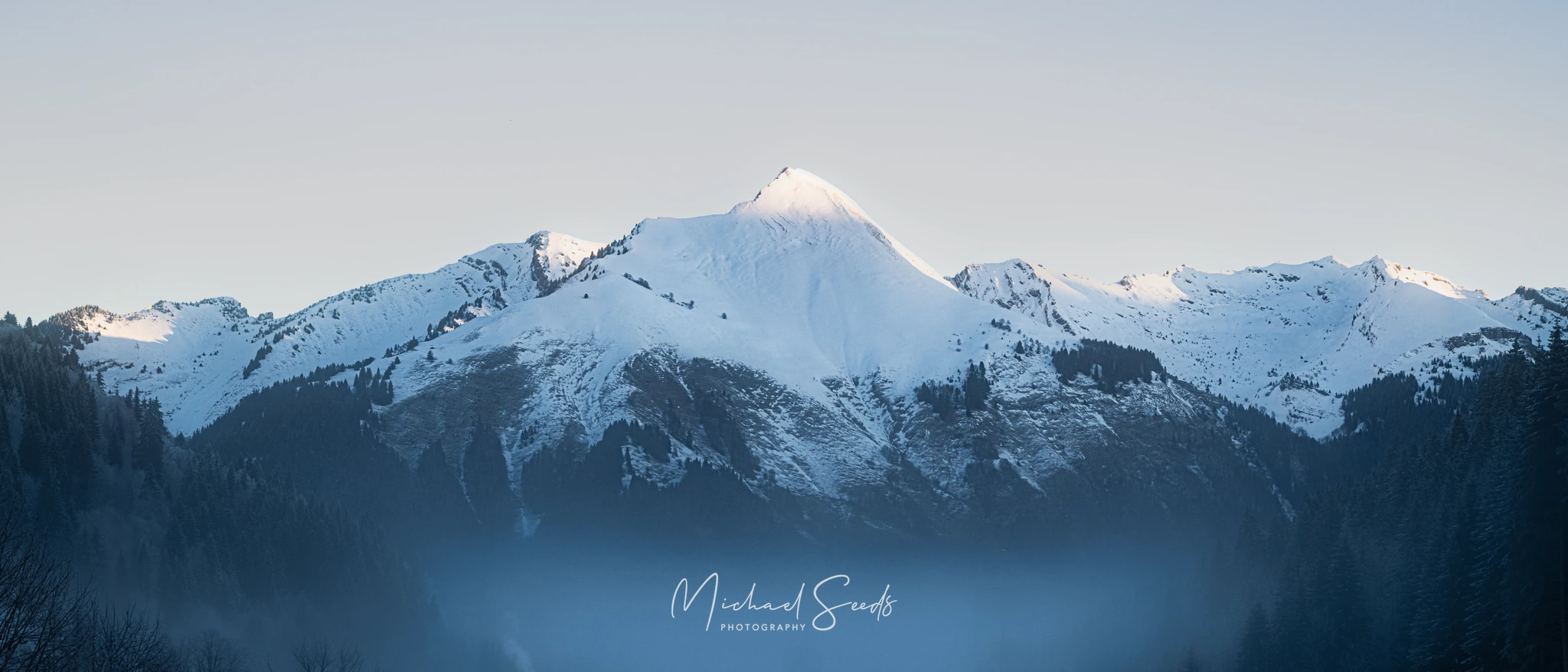 A serene winter sunrise over the Alpine peaks as seen from Les Gets, where soft morning light brushes the ridgelines and mist settles through the forested valley. A quiet moment of mountain calm.