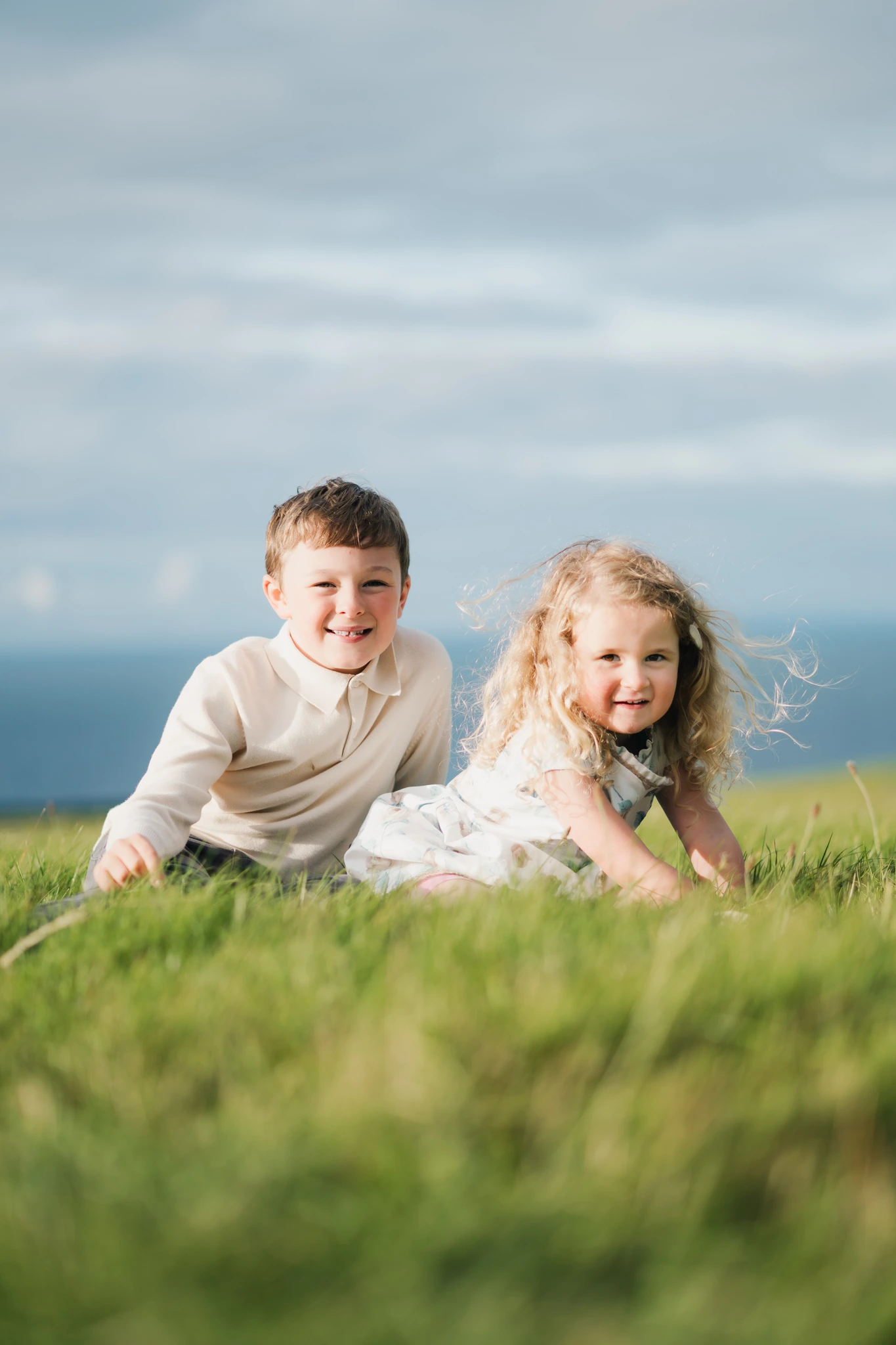 a boy and girl are sitting on a grassy field with a tree in the background