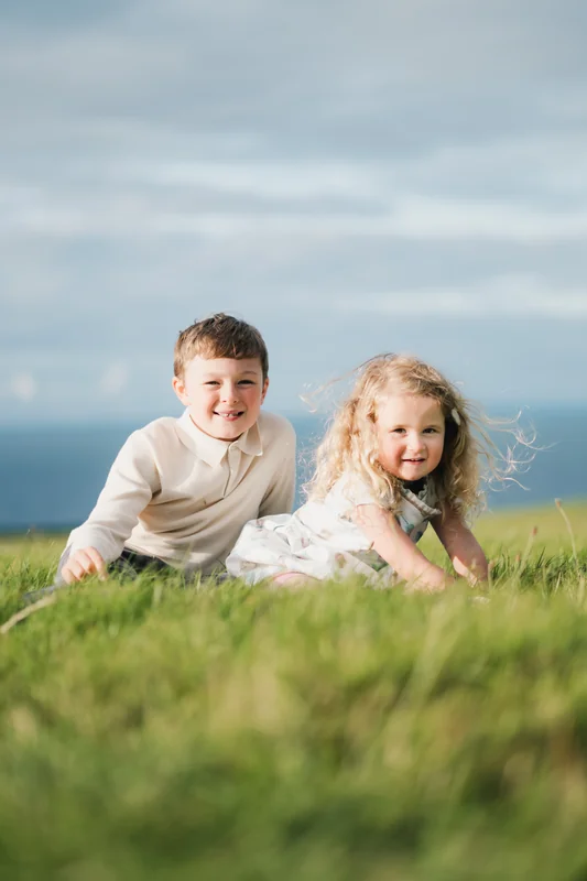 a boy and girl are sitting on a grassy field with a tree in the background