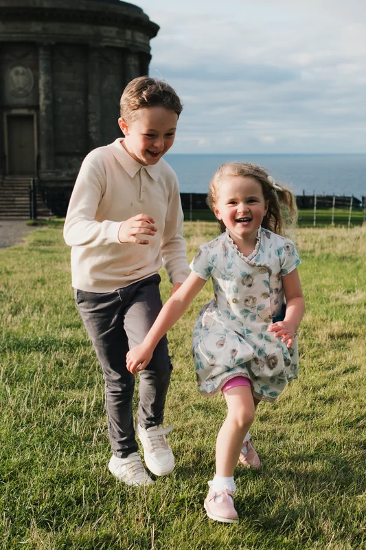 a girl and boy are running in grass near a castle