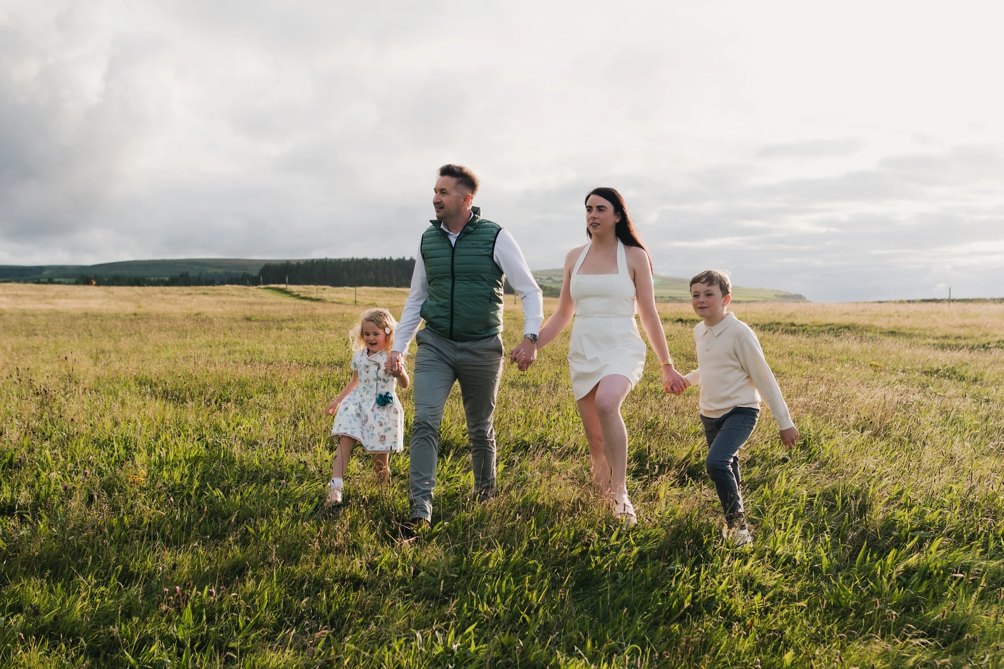a couple and their children walk through a grassy field in front of a cloudy sky