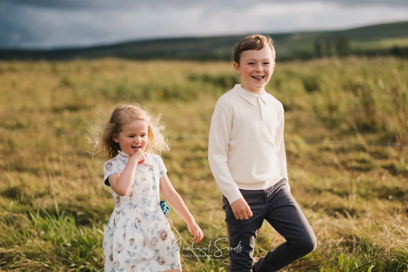 a boy and girl play in a grassy field with a cloudy sky