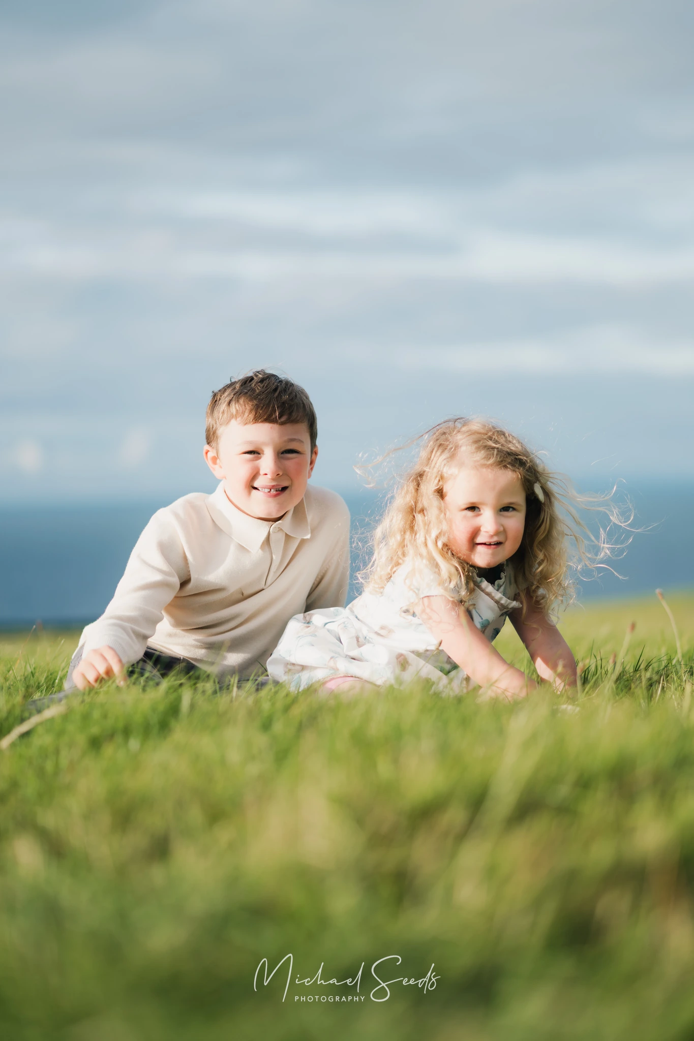 a boy and girl are sitting on the grass in front of a blue sky