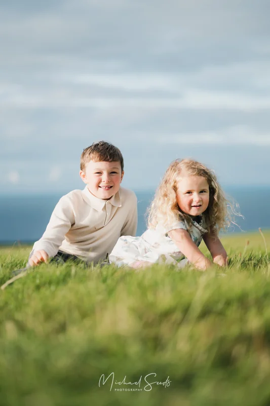 two children sit on a grassy hill in front of a blue sky