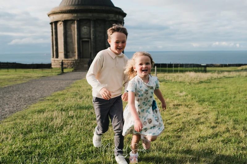 a girl and boy are running down a grassy field with a large building in the background