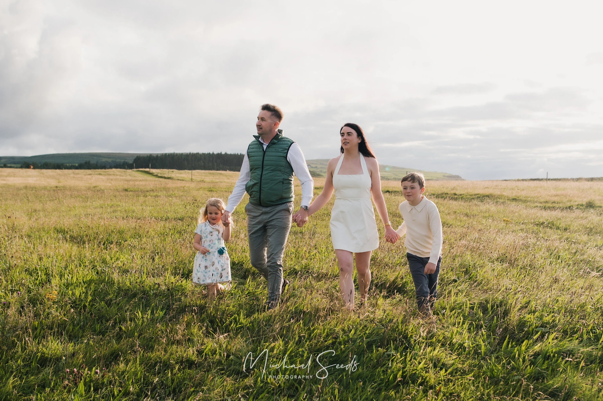 a couple and their children walk through a grassy field with a sunset behind them