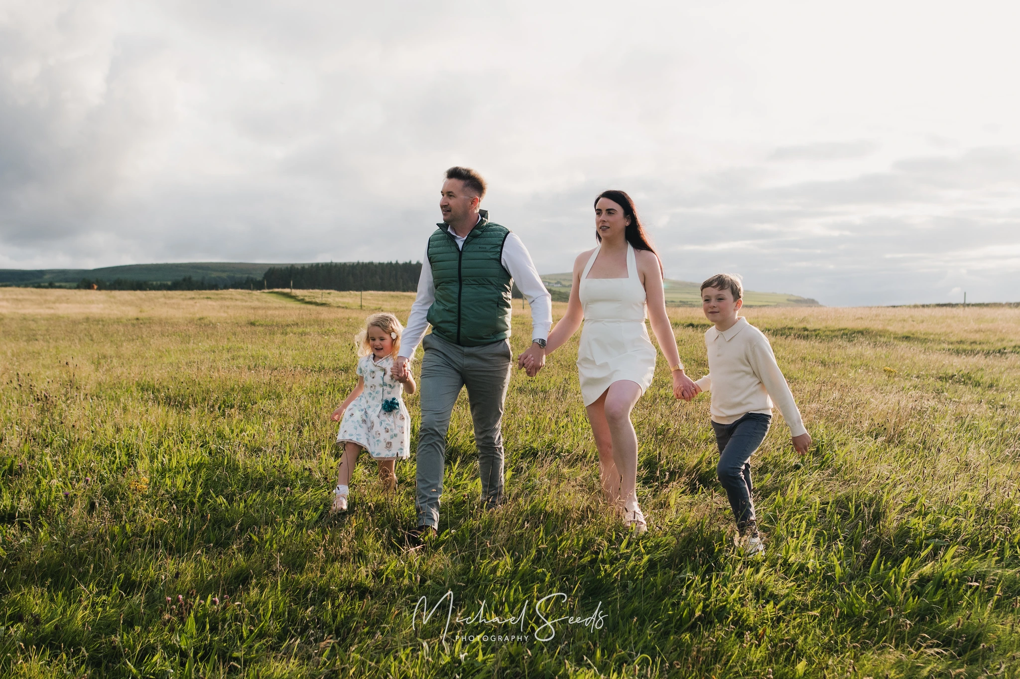 a couple and their children walk through a grassy field in front of a cloudy sky