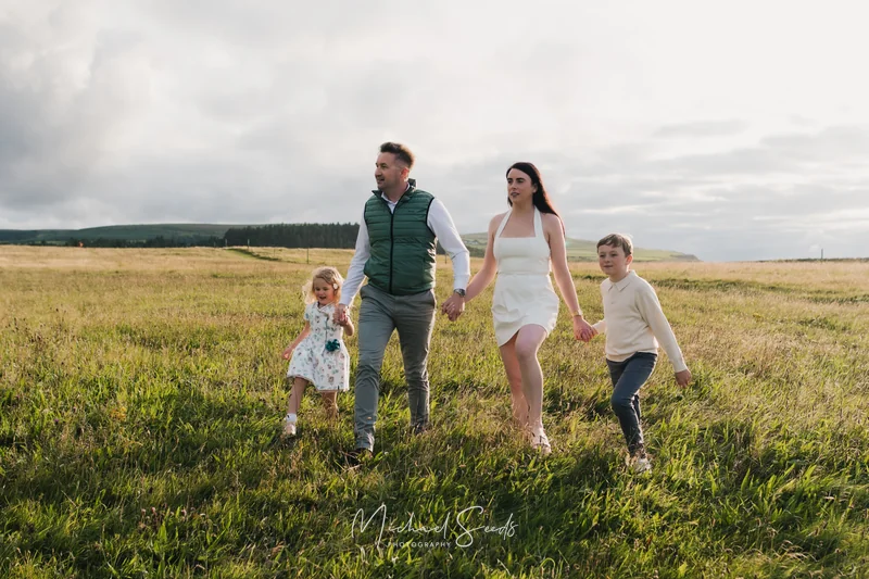 a couple and their children walk through a grassy field in front of a cloudy sky