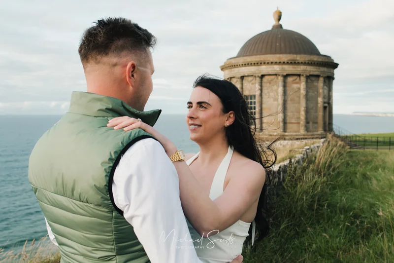 a bride and groom hugging in front of a castle overlooking the sea