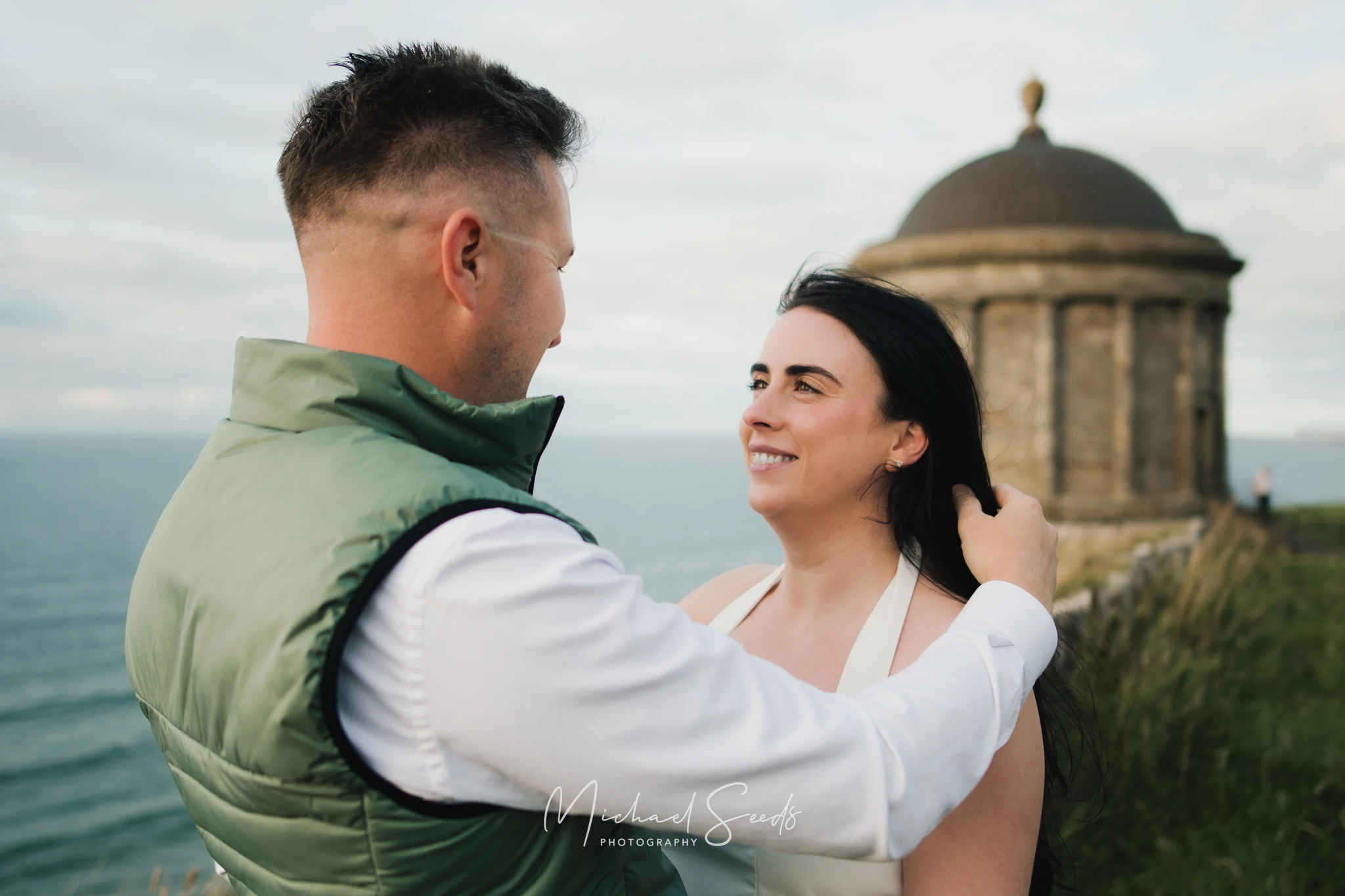 a bride and groom hugging in front of a castle overlooking the sea
