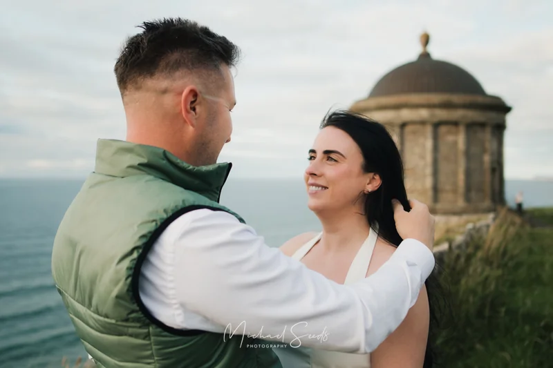 a bride and groom hugging in front of a castle overlooking the sea