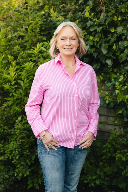 a woman wearing a pink shirt and jeans standing in front of bushes