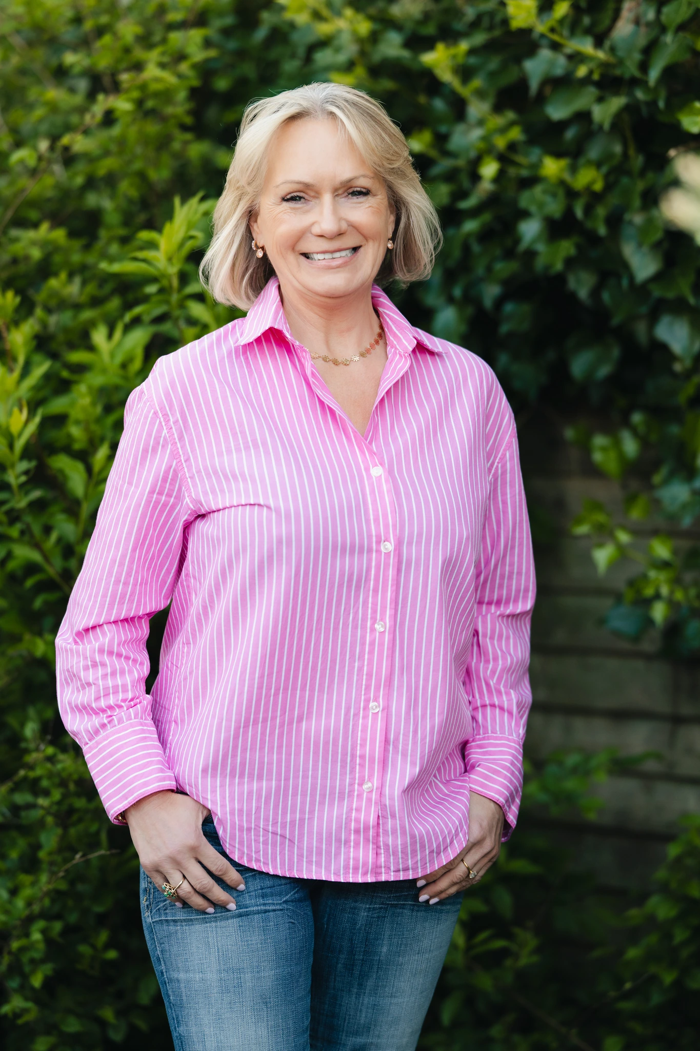 a woman in a pink shirt and jeans standing in front of bushes
