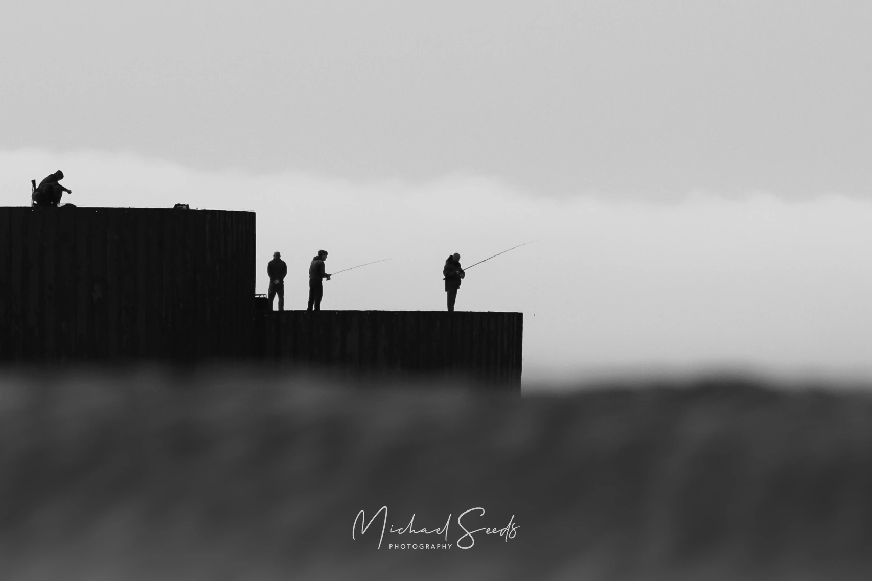 Silhouettes stand against a pale horizon, rods angled toward the sea. A quiet moment suspended between sky, water, and patience.