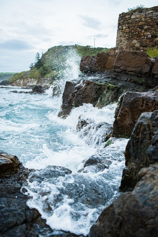 Waves crash against the rock face below the old stone wall, breaking into white swirls before retreating. The coastline holds its rugged edge, shaped continuously by the sea.