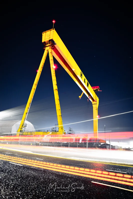 Beneath a cold Belfast sky, the great shipyard crane rises in vivid yellow, lit against the darkness. Light trails streak past at its feet, tracing modern movement beneath a monument of industrial history.