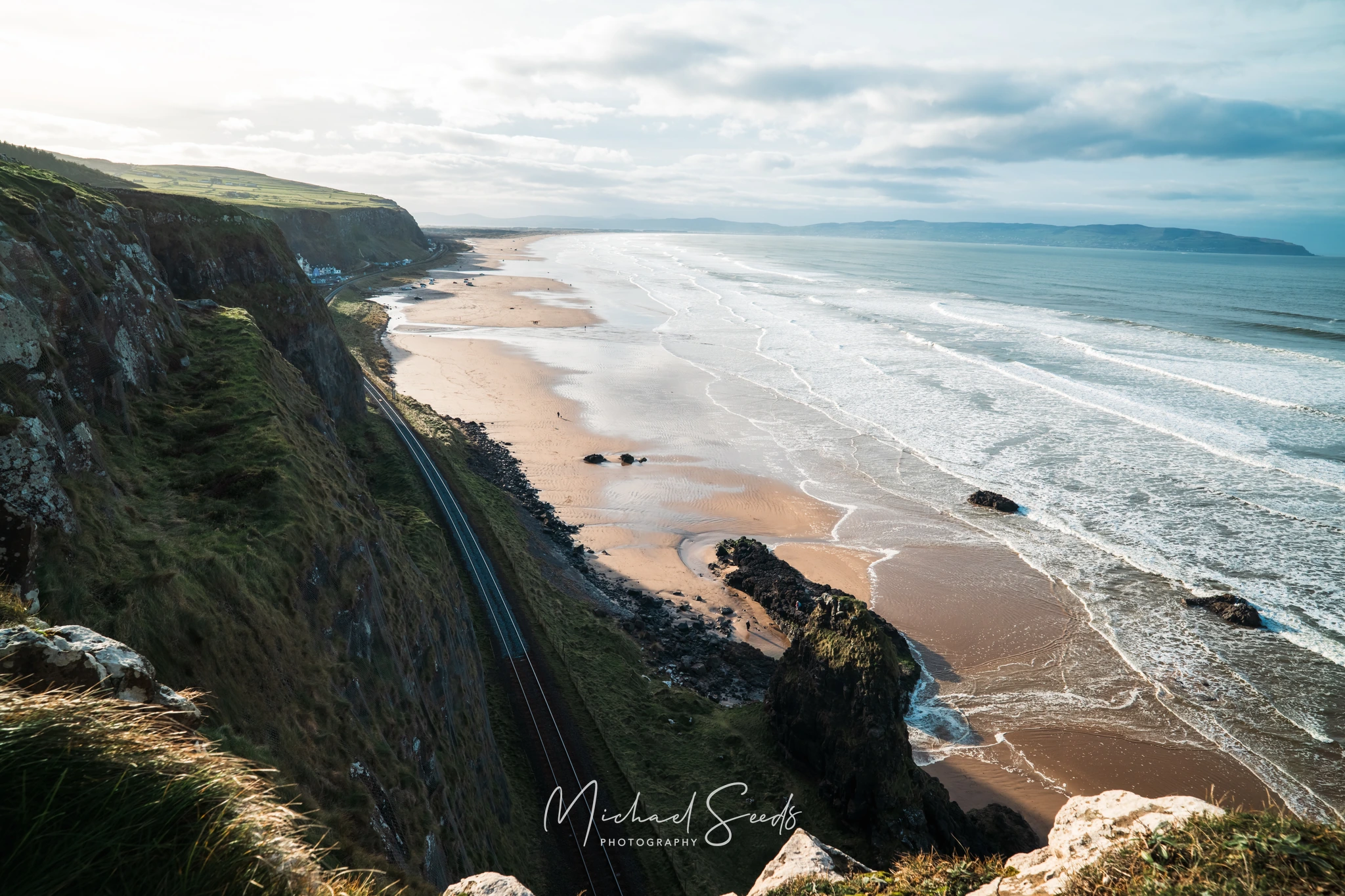 The railway hugs the cliff as the beach unfolds below, washed in silver light and rolling waves. A horizon of water and land blends into stillness—Downhill at its most expansive and calm.