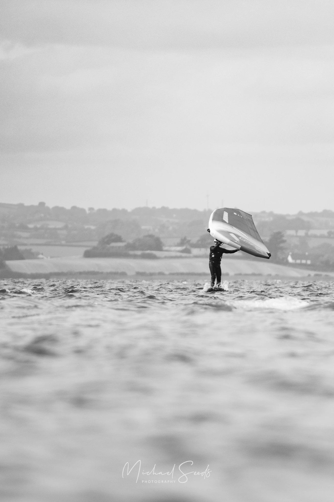 A lone rider glides above the surface, lifted by wind and foil. The blurred water foreground softens the moment into quiet movement.