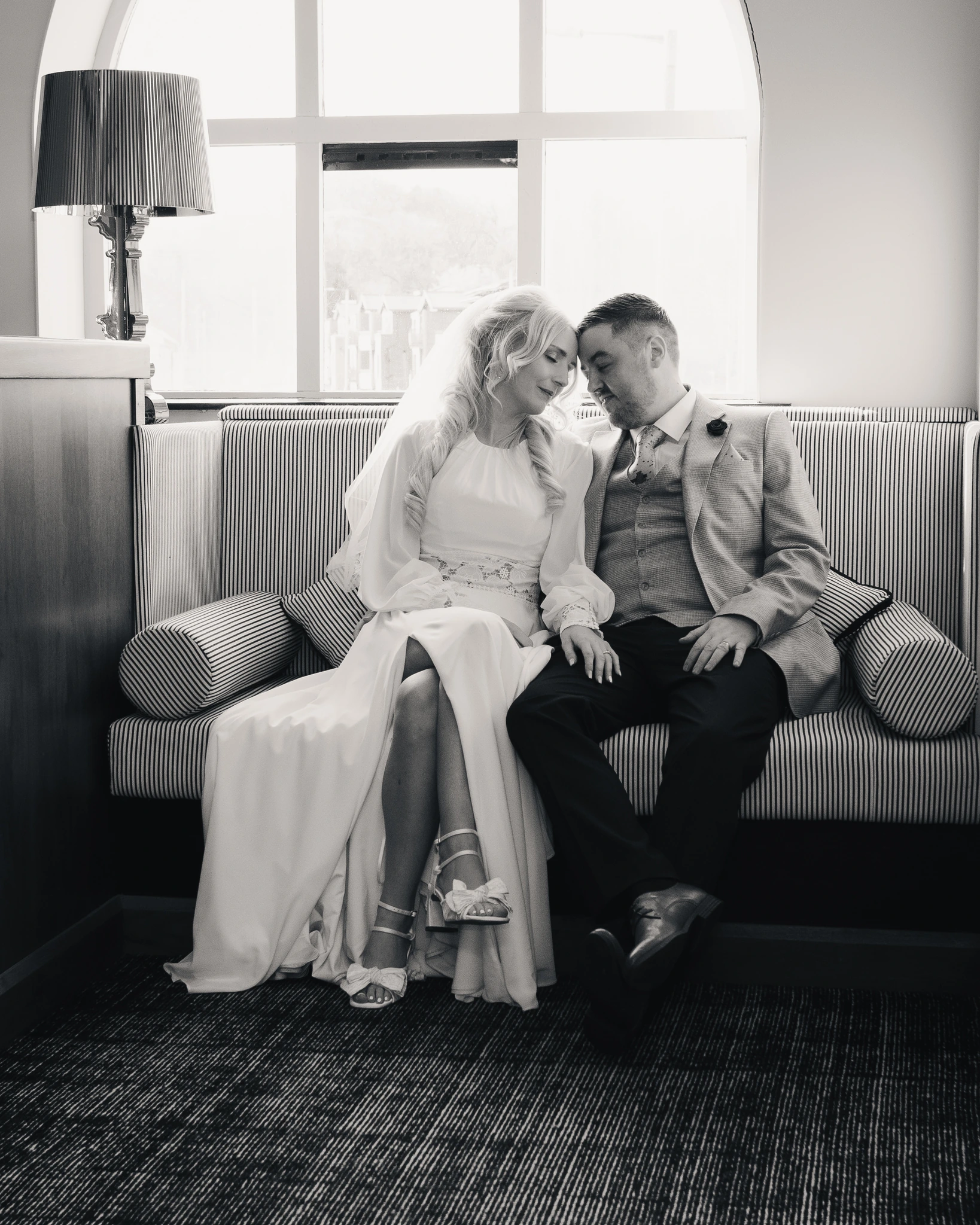 a bride and groom sit on a sofa in front of a window