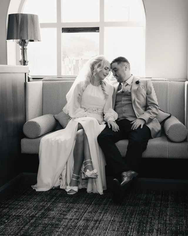 a bride and groom sit on a sofa in front of a window