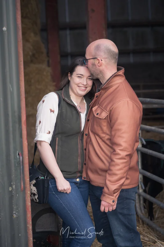 a man and woman hugging in front of a barn with sheep in the background
