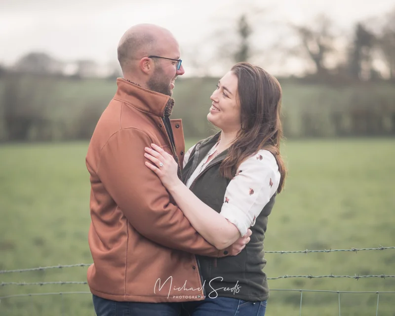 a couple hugging in front of a fence in a field