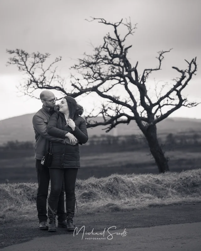 black and white image of a couple hugging a tree in a field