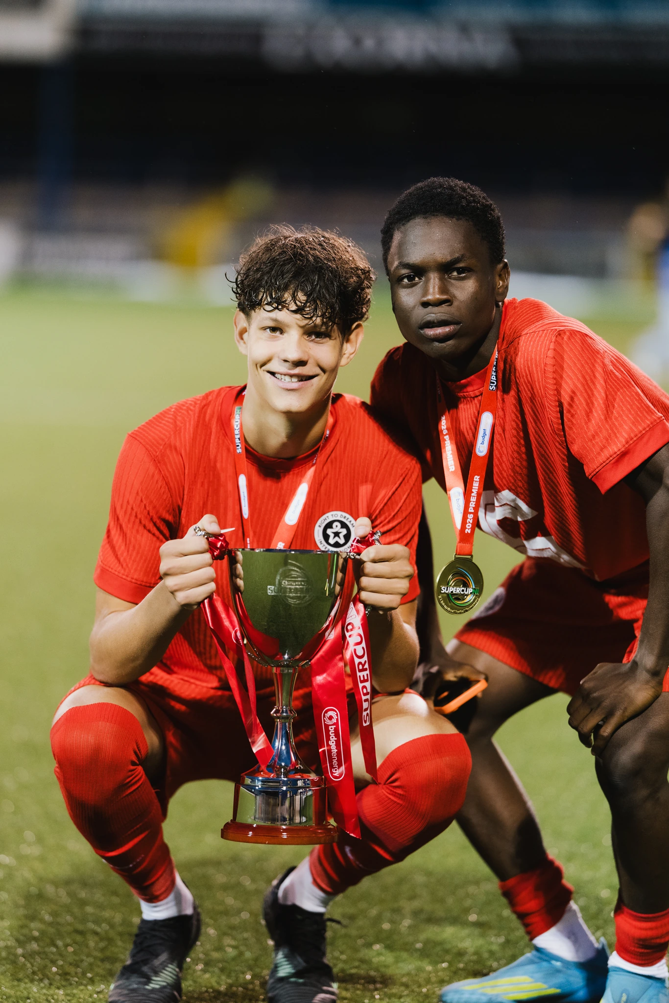 two young men pose with a trophy in front of a soccer field