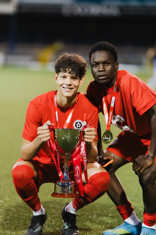two young men pose with a trophy in front of a soccer field