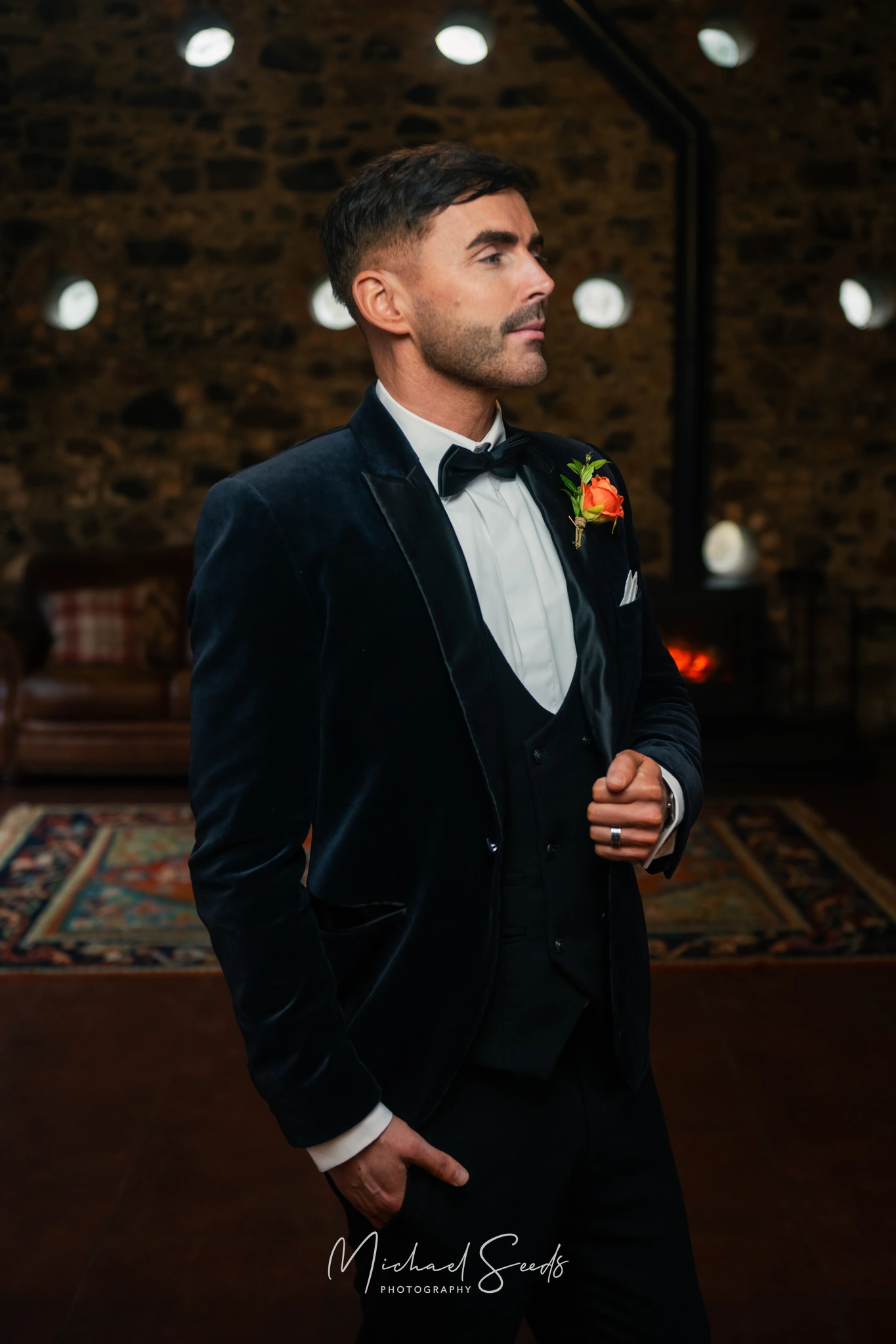 a man in a tuxedo standing in front of a fireplace