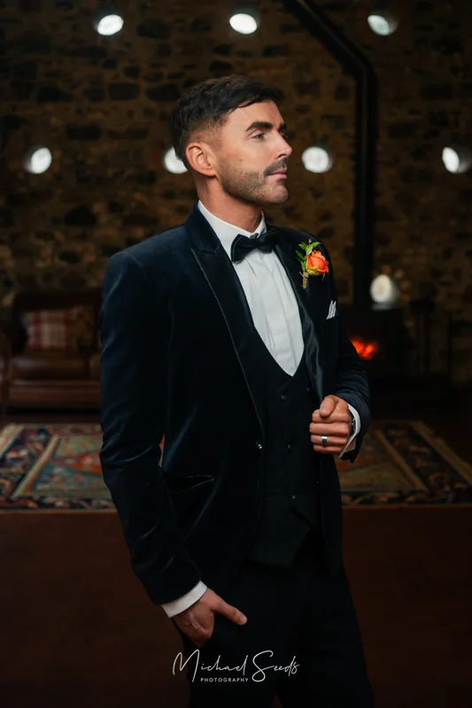 a man in a tuxedo standing in front of a fireplace