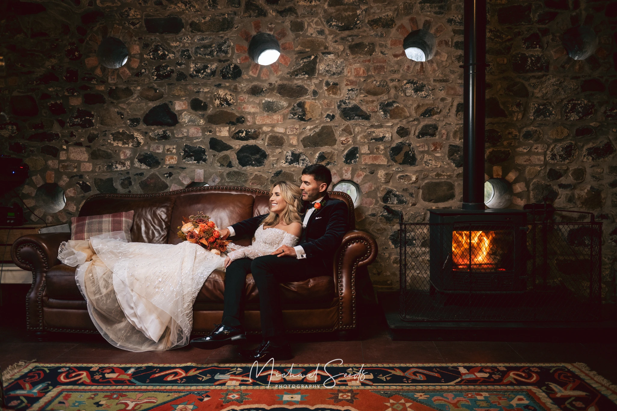 a bride and groom sit on a sofa in front of a fireplace