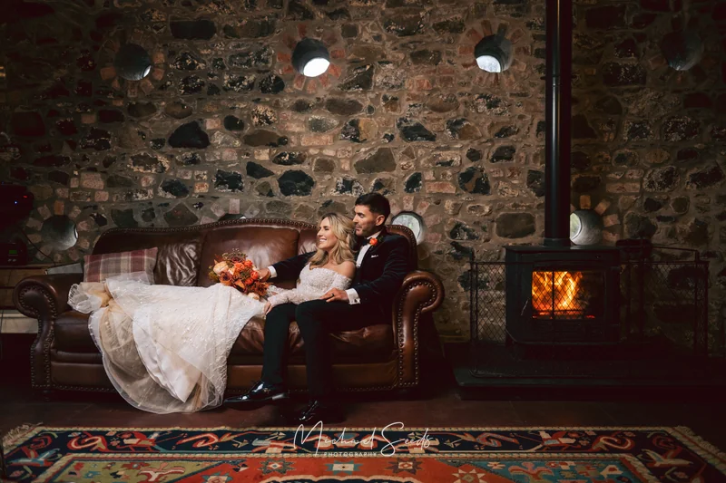 a bride and groom sit on a sofa in front of a fireplace