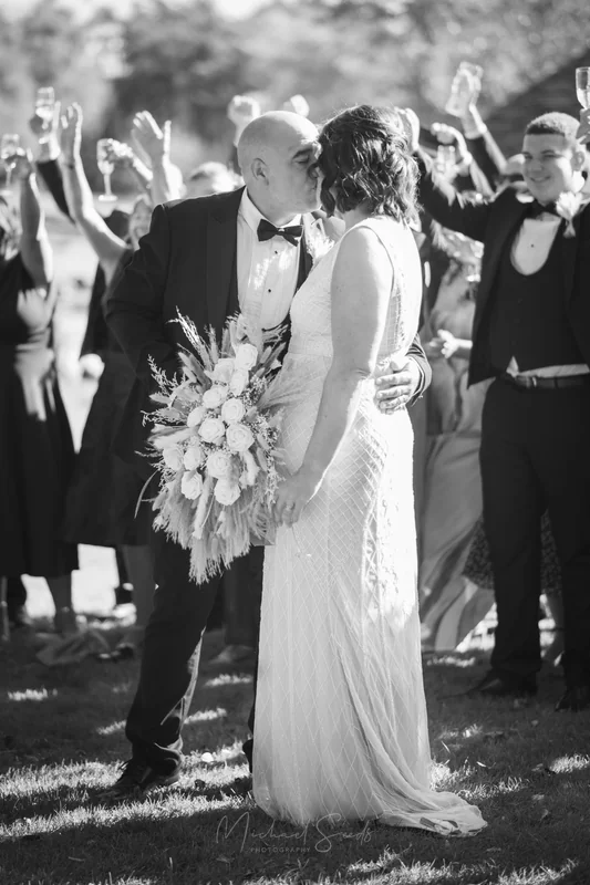 Bride and groom kissing outside with guests cheering behind them at a Belfast wedding.