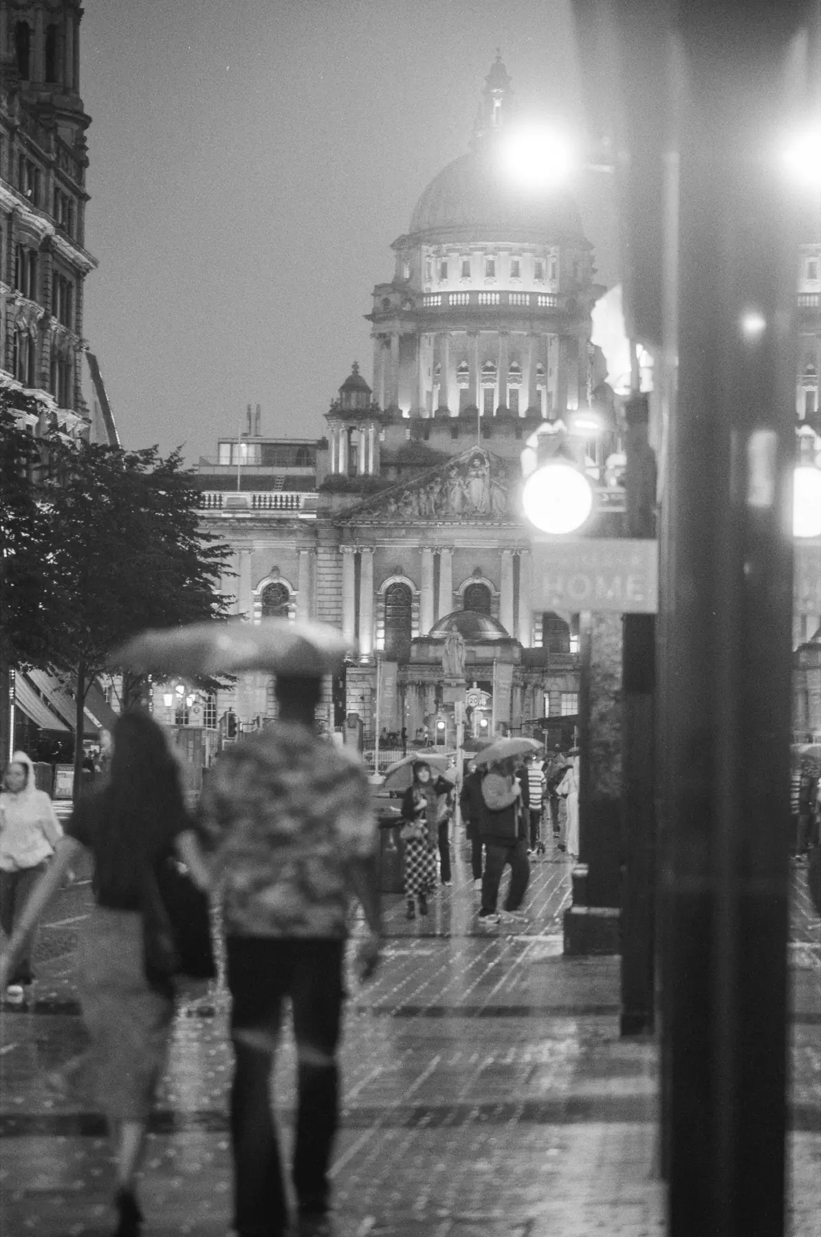 People walking through rain near Belfast City Hall at night.